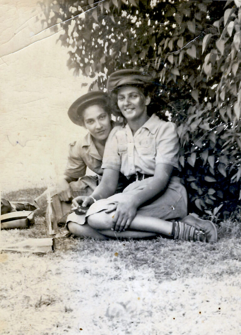 Grey and white photo of two women in solider uniform sitting on grassy ground in front of leafy bush.