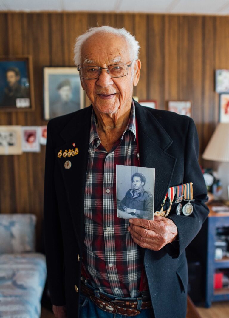 Colour photo of older man in formal suit jacket, military medals affixed, holding up grey and white photo.